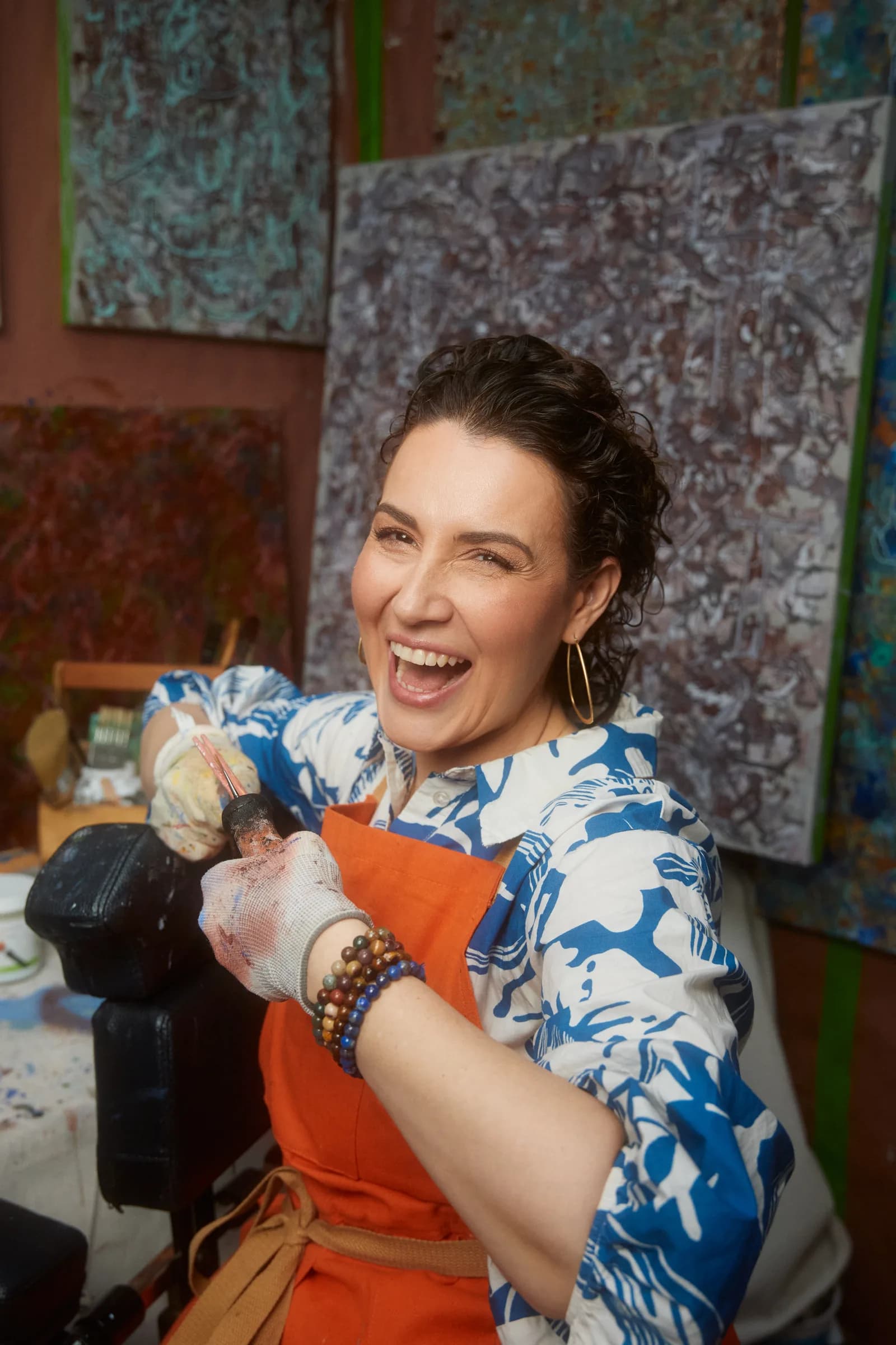 Ana Violeta Horta laughing in her studio, wearing an orange apron with paint on her hands, surrounded by her paintings